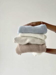 stack of neutral folded sweaters in beige, white, and light blue held by hand